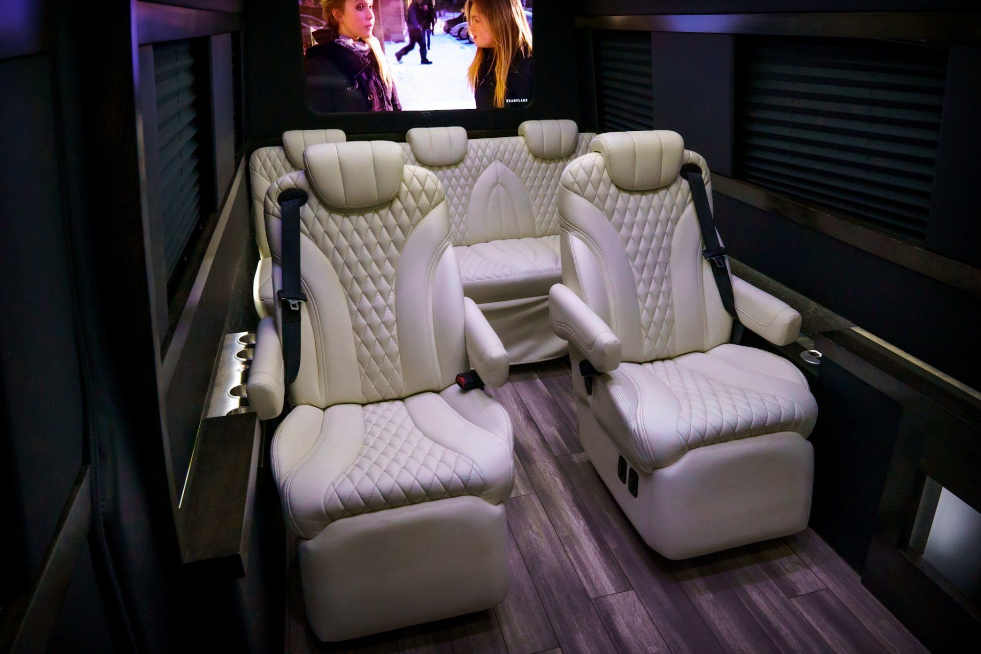 Interior view of a luxury van with white leather seats, a TV screen, and wood-look flooring.