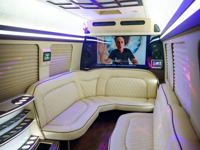 Interior of a luxury van with white leather seating, large TV screen, and ambient lighting.