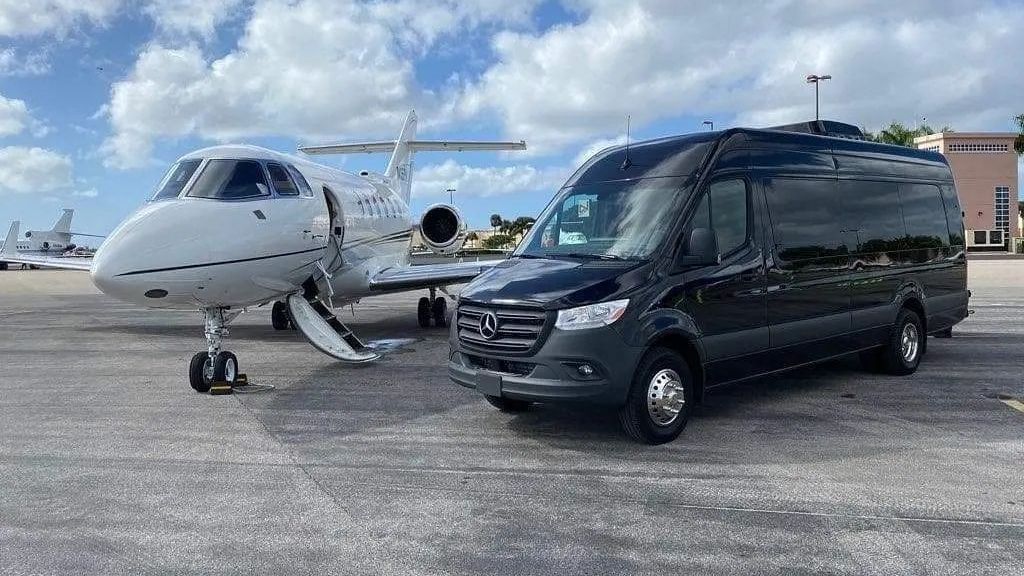 Black van parked next to a private jet on a tarmac under a cloudy sky.