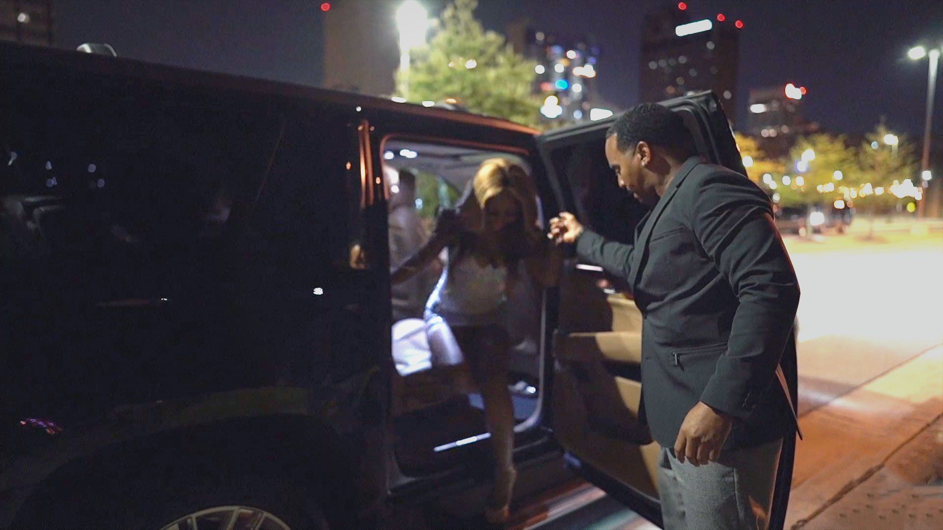 Man helps person exit a dark SUV at night; city lights in background.