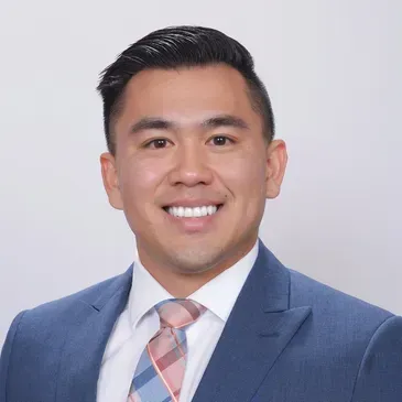 Studio headshot of a smiling man in a blue suit and pink striped tie on a light background
