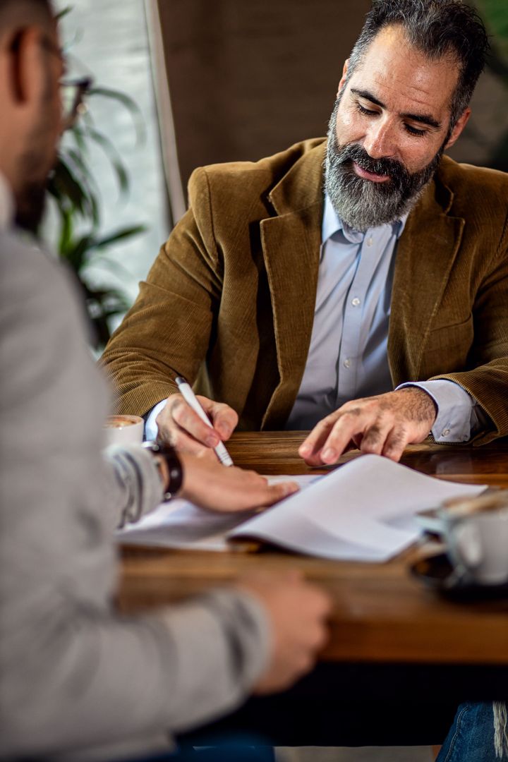 Man in brown blazer reviewing documents and writing at a table during a meeting