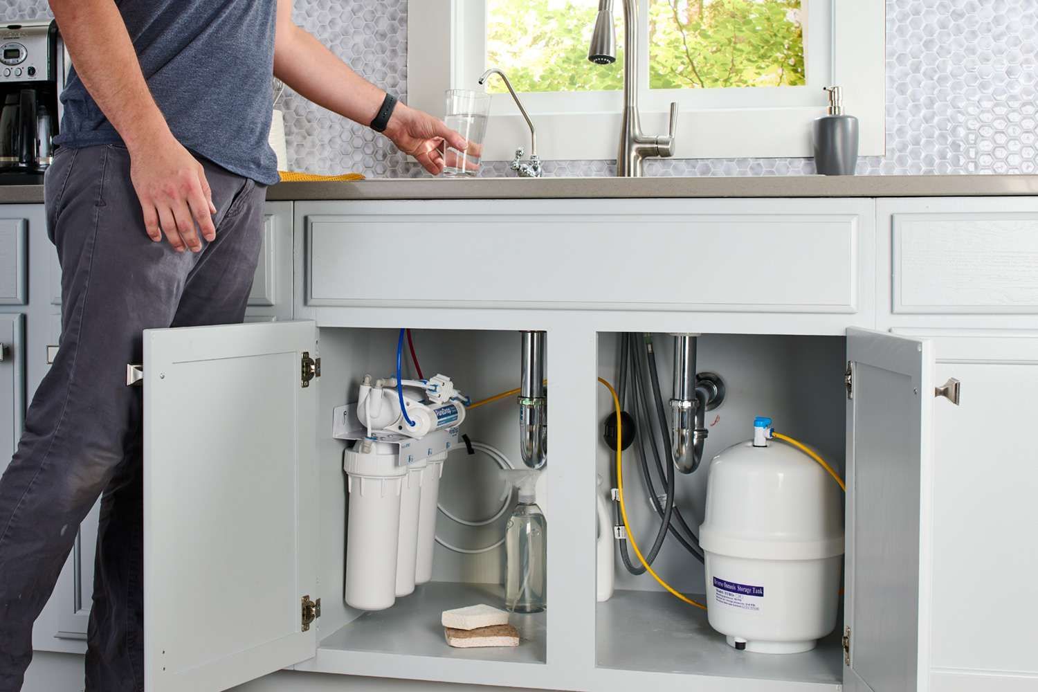 Person at a kitchen sink with open cabinets showing plumbing and water filtration equipment