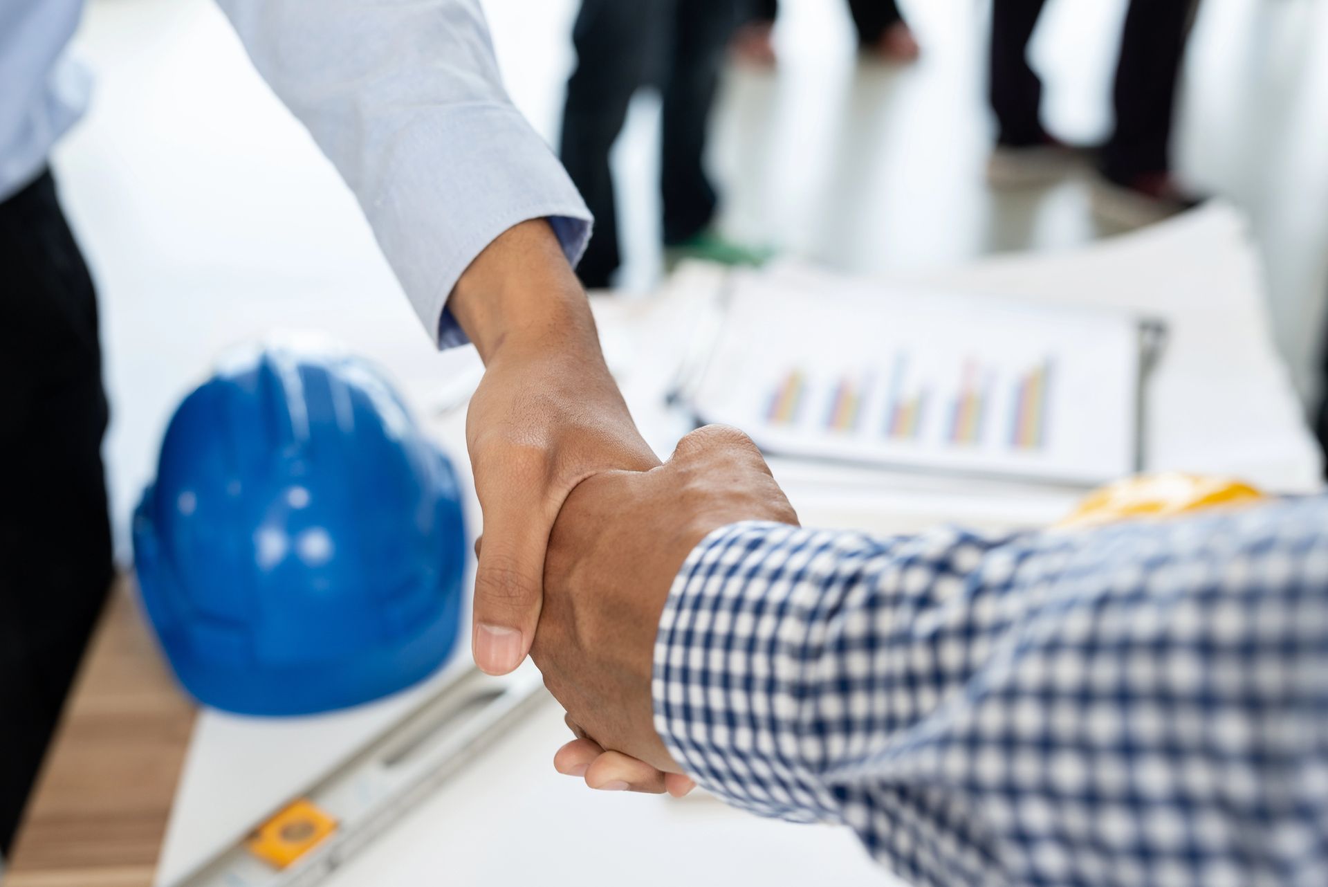 Due persone si stringono la mano su un tavolo con un progetto, un casco e degli attrezzi.