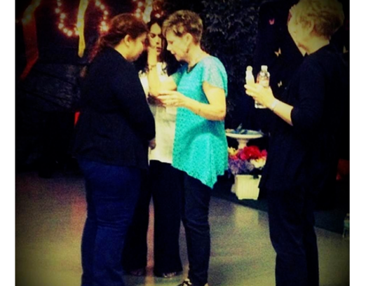 People standing and talking indoors, one in a turquoise top holding a drink, others in dark clothing.