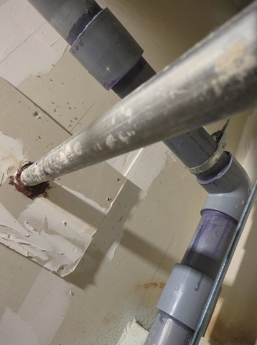 Close-up of metal pipes and gray PVC plumbing against a beige wall, with a rusty joint.