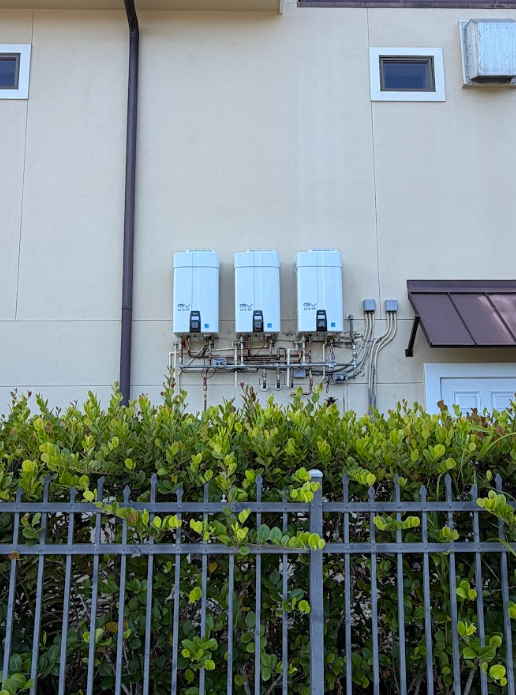 Row of white utility boxes on a beige building above a hedge and metal fence