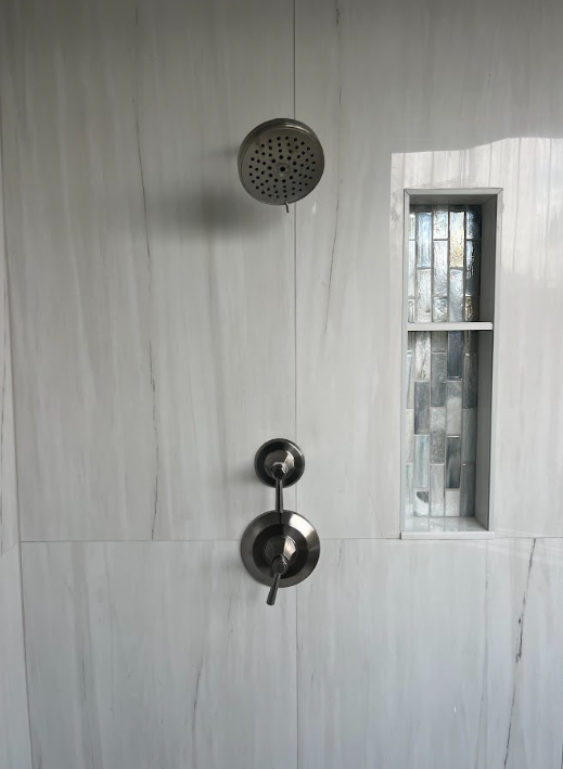 Shower wall with chrome controls, round showerhead, and narrow window beside it