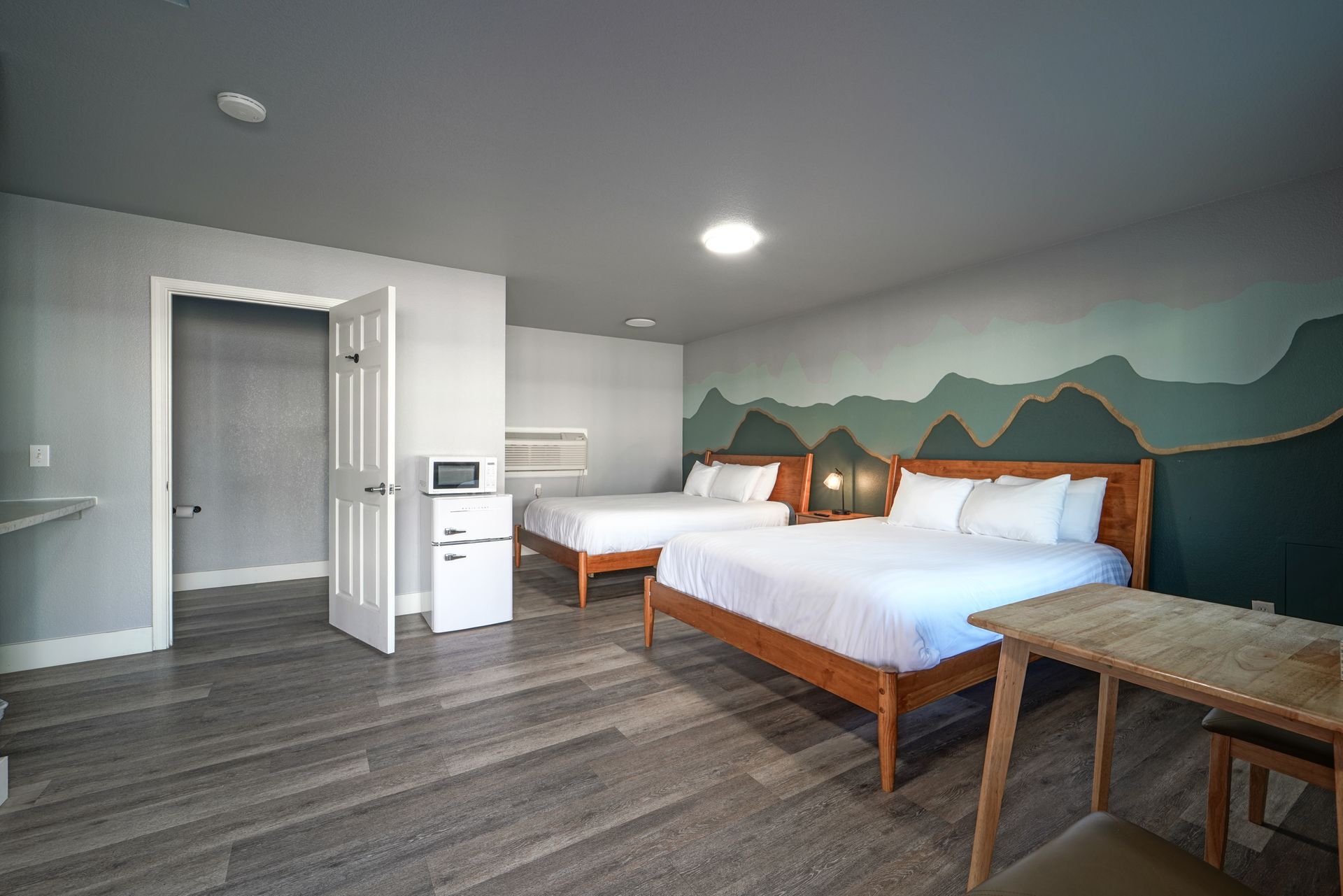 Spacious hotel room with two beds, mural wall, gray floors, and small table near the window.