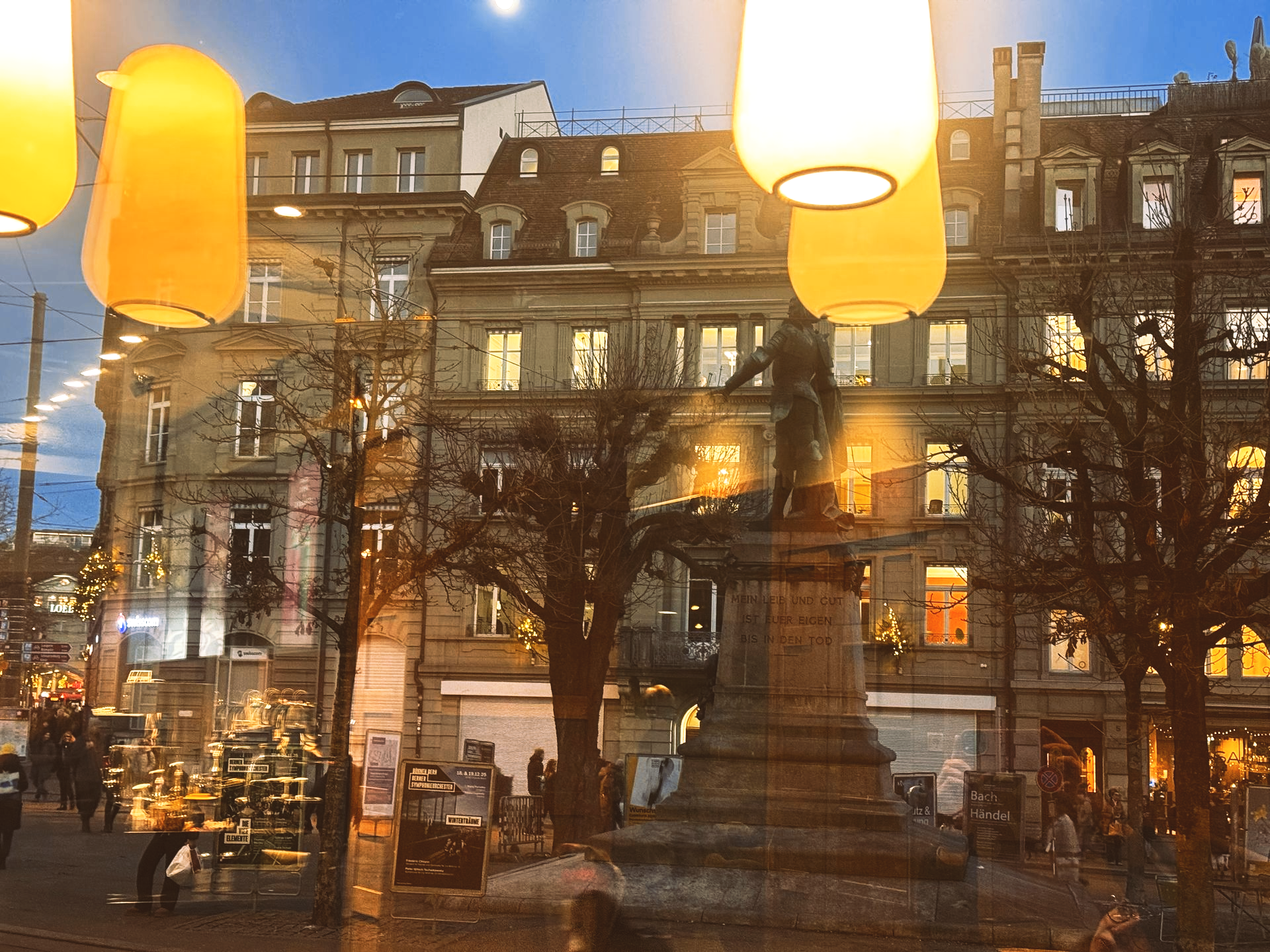 Stadtplatz am Abend mit einem Steinmonument, beleuchteten Gebäuden, kahlen Bäumen und hängenden Laternen