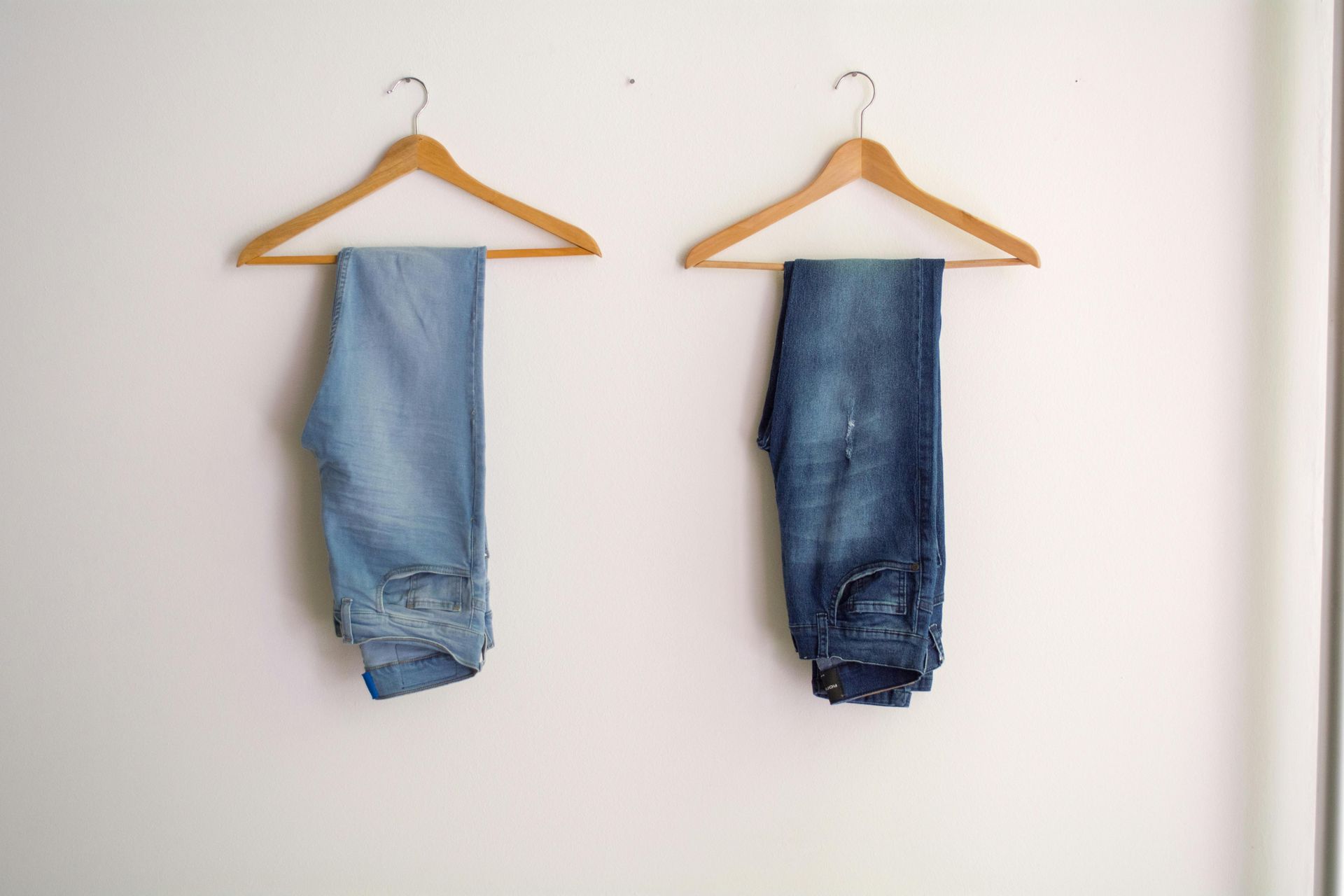 Zwei Jeans hängen auf einem hölzernen Kleiderbügel an einer weißen Wand.