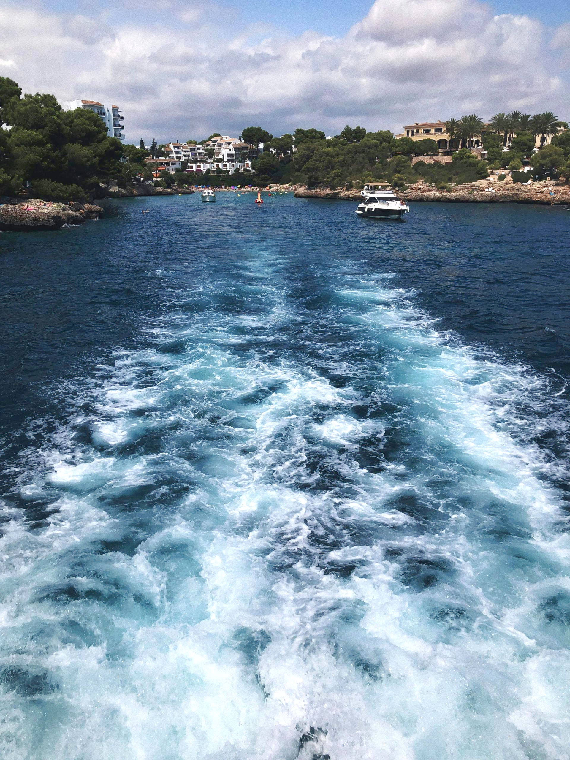 Bootswellen auf blauem Wasser nahe einer baumbestandenen Küste und Häusern unter einem bewölkten Himmel