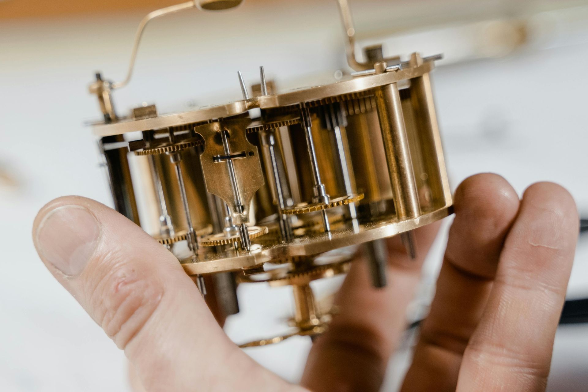 Nahaufnahme von Händen, die ein kleines mechanisches Gerät aus Messing mit Zahnrädern und Zylindern halten.