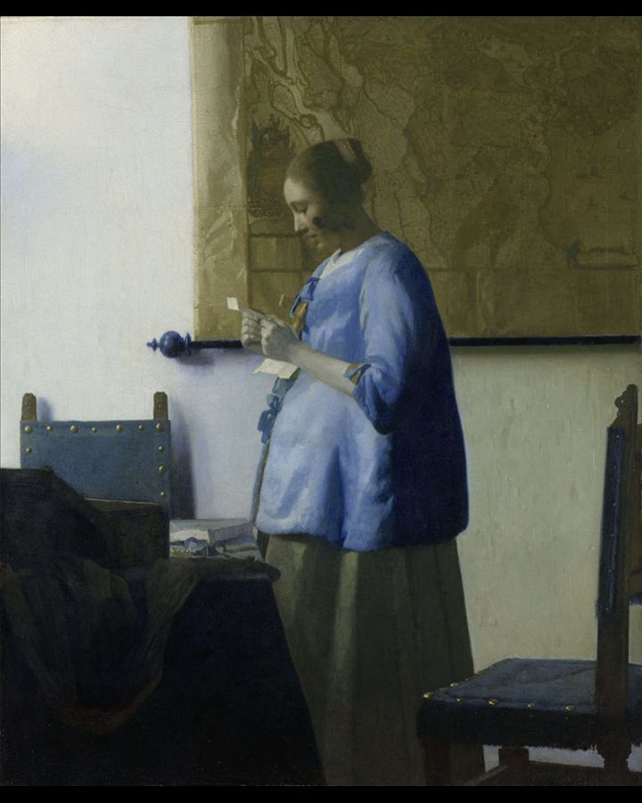 Vrouw in blauwe jas leest een brief in een kamer. Natuurlijk licht, eenvoudige meubels, een schilderij aan de muur.