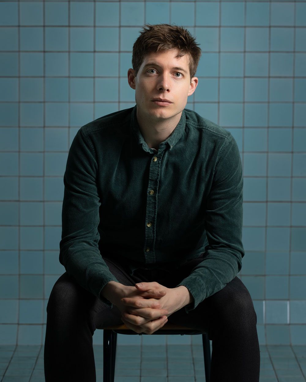Een man in een turquoise shirt zit met zijn handen ineengevouwen voor een muur van blauwe tegels.