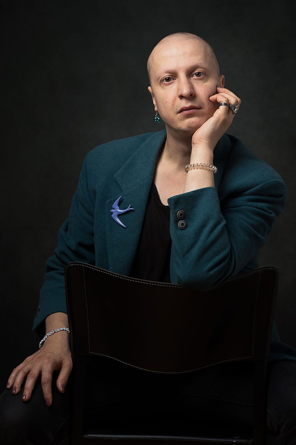 Een kale persoon zit, met de kin op de hand. Hij draagt ​​een turquoise jas, een zwart shirt en sieraden. 