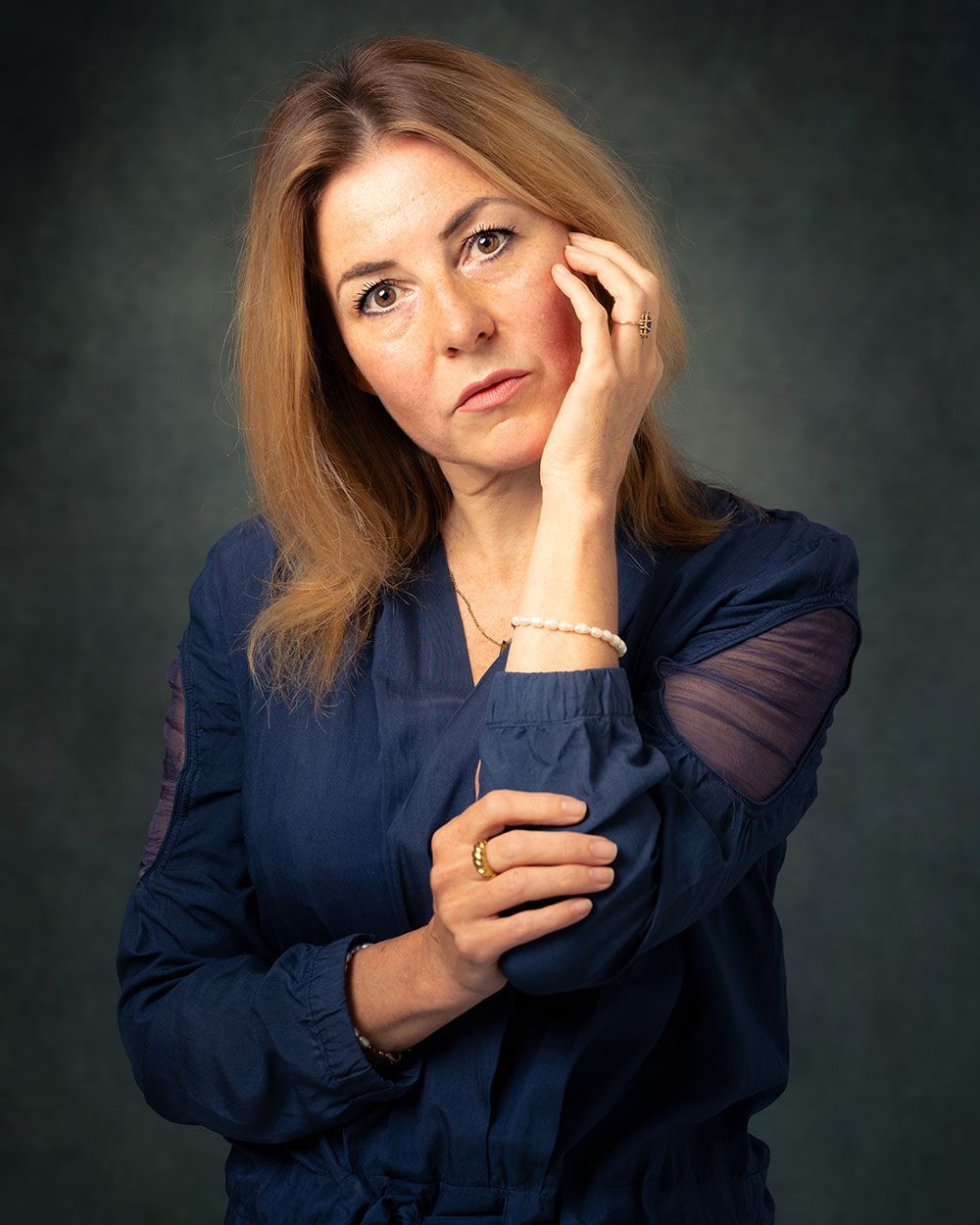 Vrouw in een blauwe blouse, met haar hoofd in haar hand, kijkt de kijker aan tegen een donkergroene achtergrond.