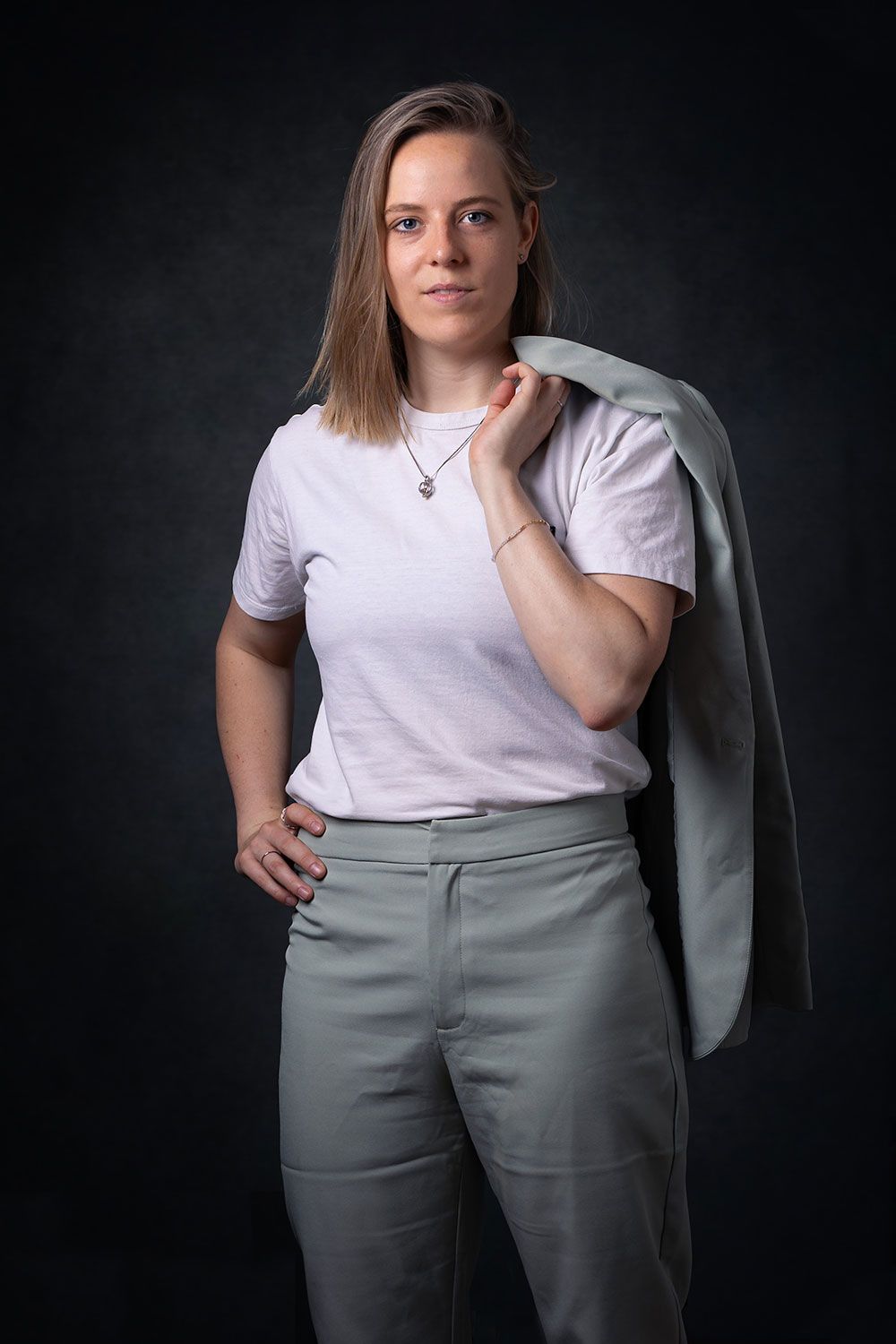 Vrouw in een lichtgekleurde broek en T-shirt, blazer over de schouder, hand in de zij, staand tegen een donkere achtergrond.