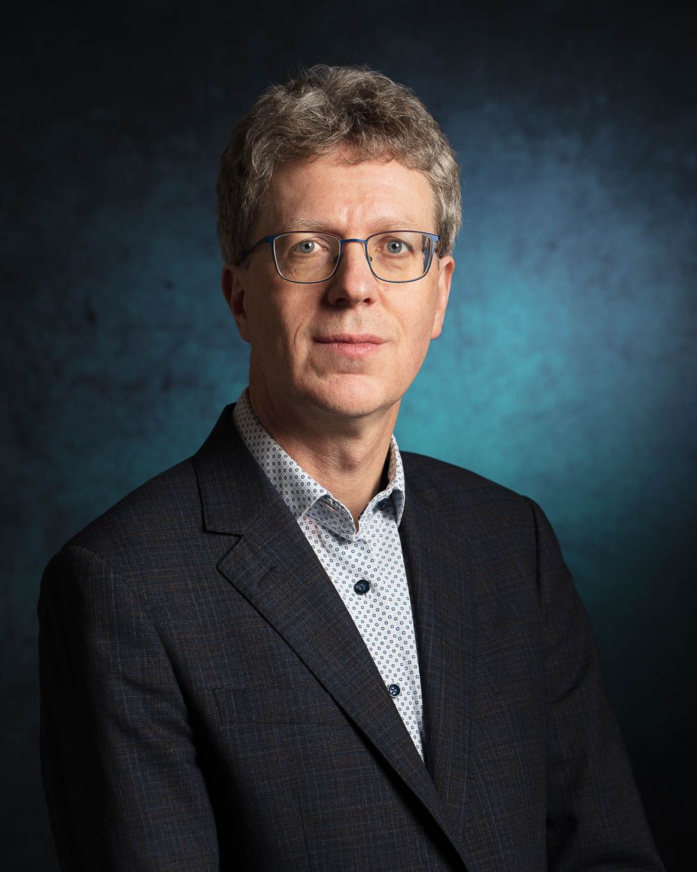 Een man met bril en colbert kijkt de kijker aan tegen een donkerblauwe achtergrond.