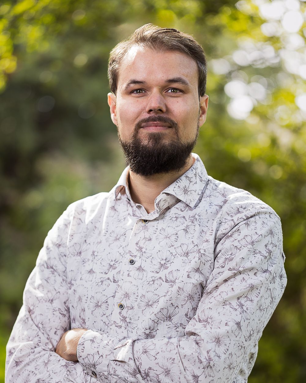 Een man met een baard en een overhemd met een patroon, met de armen over elkaar, staat buiten voor een groenperceel.