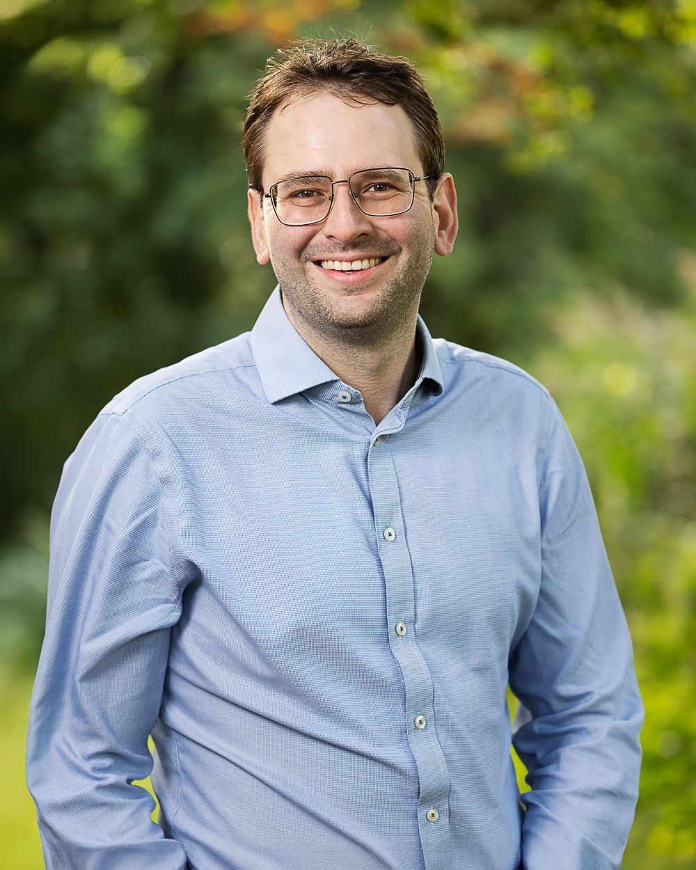 Een man met een bril glimlacht in een blauw overhemd, buiten tegen een groene, wazige achtergrond.