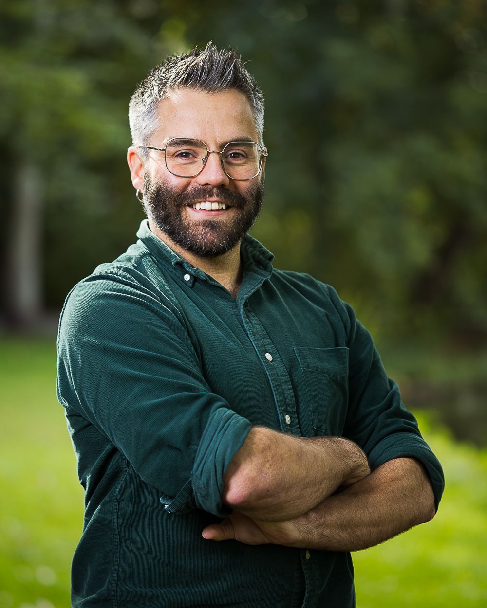 Man met bril en baard, armen over elkaar, glimlachend. Draagt ​​een groen corduroy shirt, buiten op het gras.