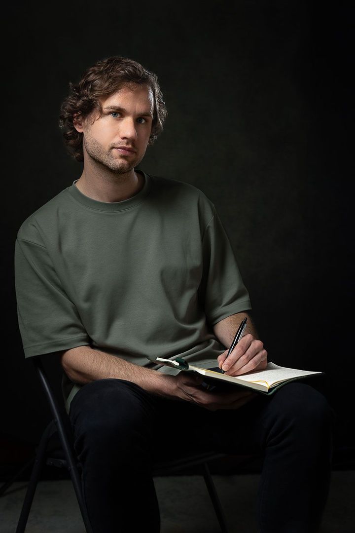 Man in groen shirt, zittend op een stoel, schrijft in een notitieboekje. Donkere achtergrond.