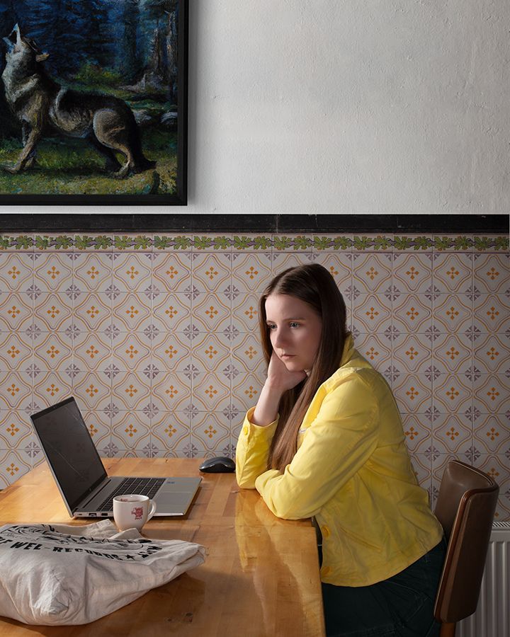 Vrouw in gele jas werkt op haar laptop aan een tafel en kijkt peinzend.