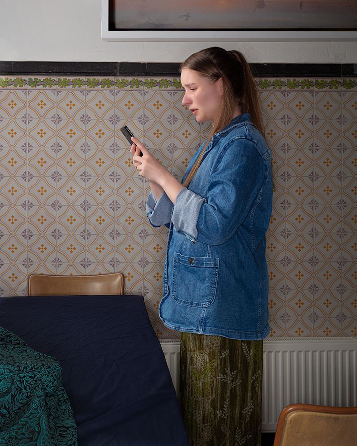 Een vrouw in een spijkerjasje kijkt op haar telefoon, staand naast een blauw matras en een muur met een patroon.
