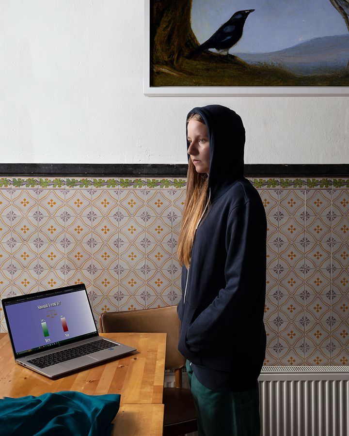 Vrouw in hoodie kijkt naar een laptop op een houten tafel, met op de achtergrond een betegelde muur en daarboven een schilderij van een vogel.
