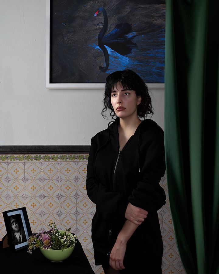 Een vrouw in een zwarte hoodie staat bij een tafel met een foto, bloemen en een schilderij van een zwarte zwaan.