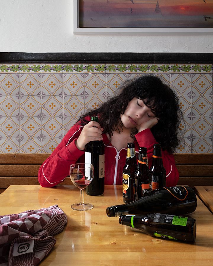 Een vrouw ligt te slapen aan een tafel met lege flessen en een glas wijn, in een bar.