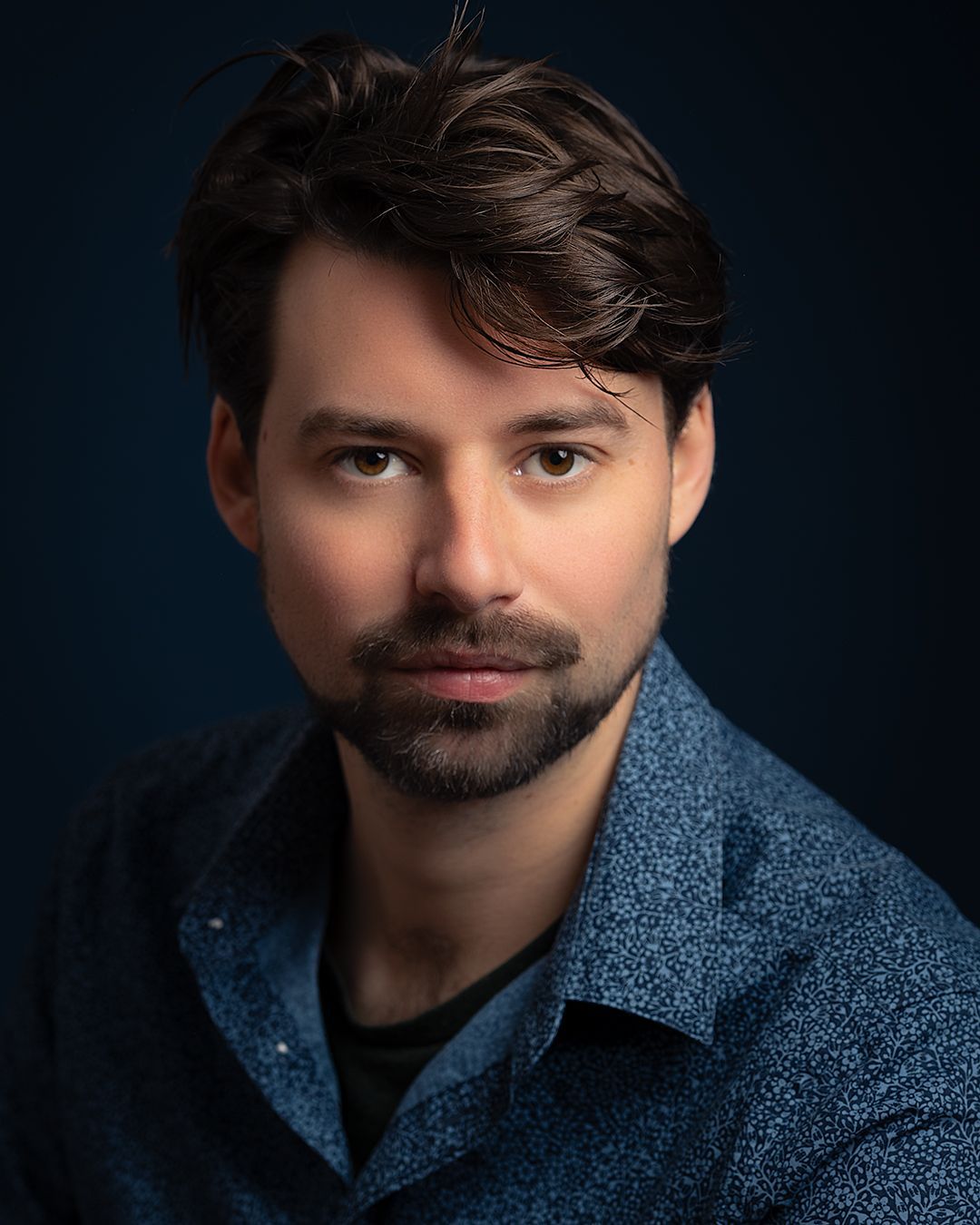 Man met bruin haar en een baard, gekleed in een blauw overhemd, kijkt in de camera. Donkerblauwe achtergrond.