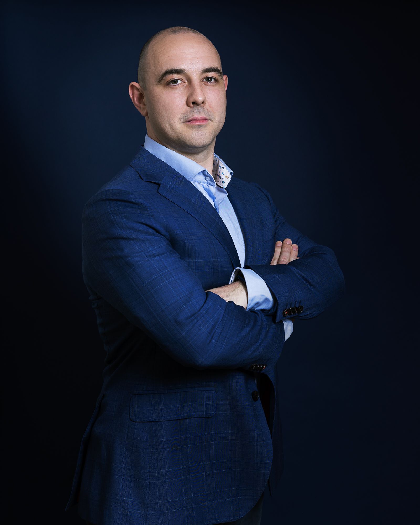 Een man in een blauwe blazer met de armen over elkaar, staand tegen een donkerblauwe achtergrond.