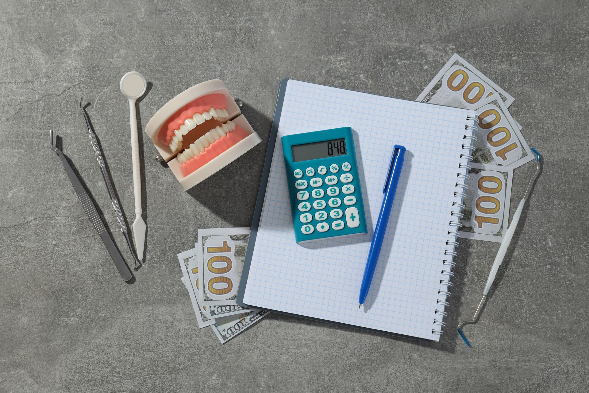 Dental tools, teeth model, calculator, notebook, pen, and money on a gray surface, possibly related to dental costs.