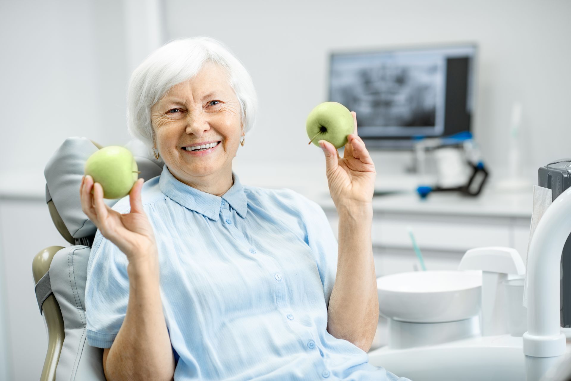 A smiling person in a dentist's office chair holds a green apple in each hand.