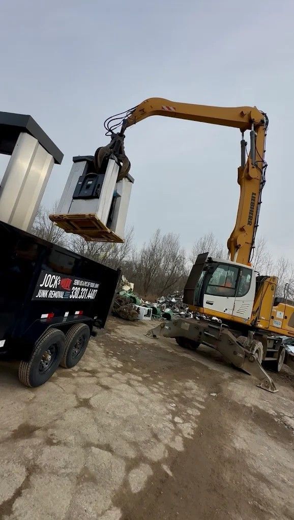 Yellow excavator lifting a metal dumpster over a black truck in a muddy lot