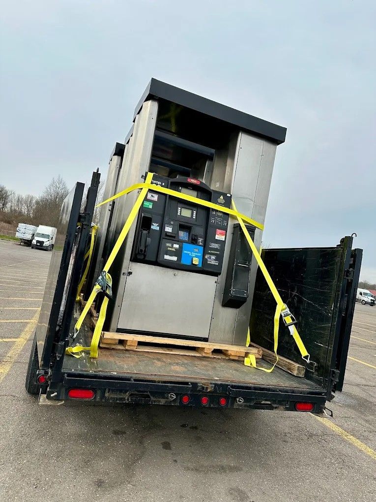 Large machine strapped with yellow tie-downs on a flatbed trailer in a parking lot.