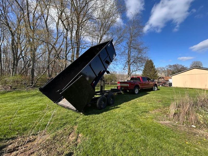 Large black dump trailer tipped up on a grassy slope beside a red pickup and beige building under blue sky