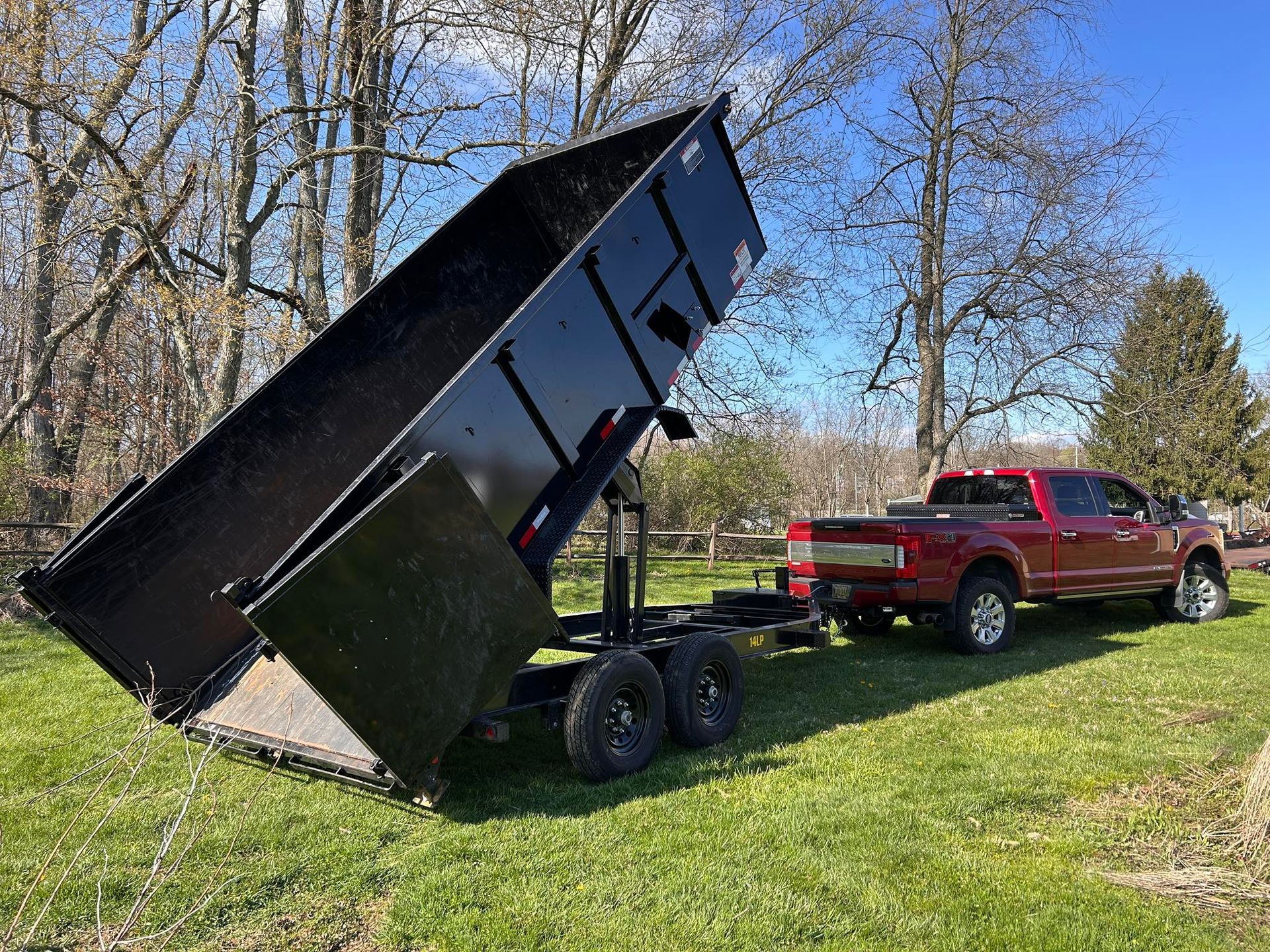 A red pickup truck dumping a black trailer bed on a grassy field near trees.