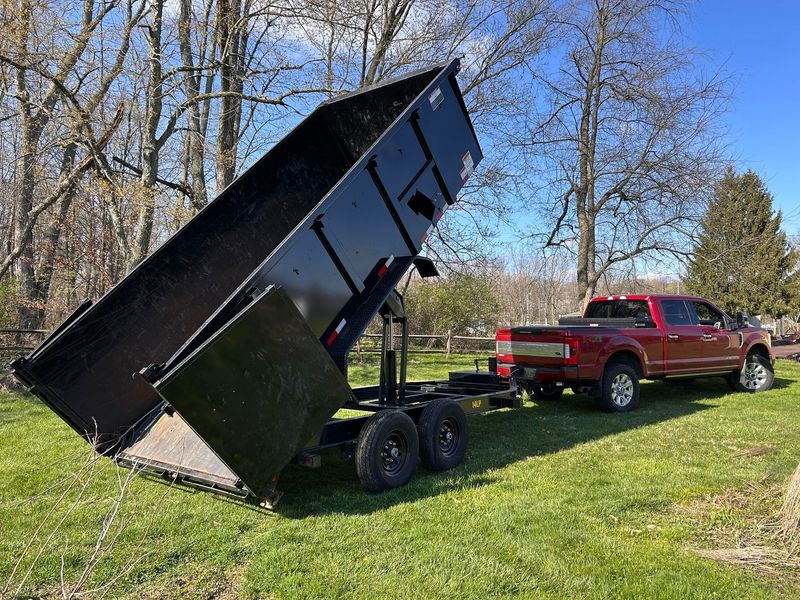A red pickup truck dumping a black trailer bed on a grassy field near trees.