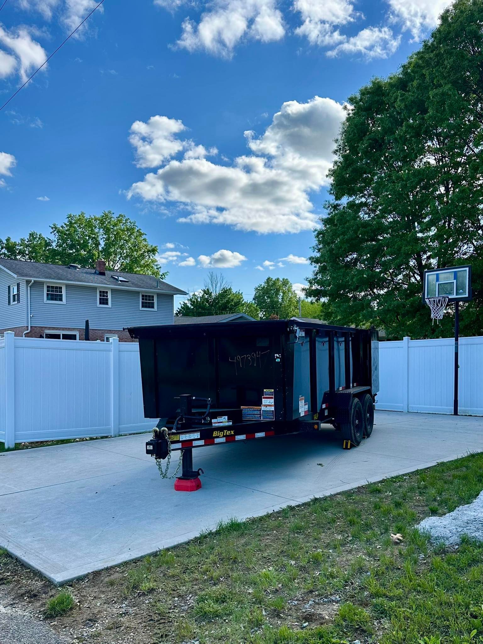 Black utility trailer parked on a concrete driveway beside a white fence and basketball hoop under a blue sky.