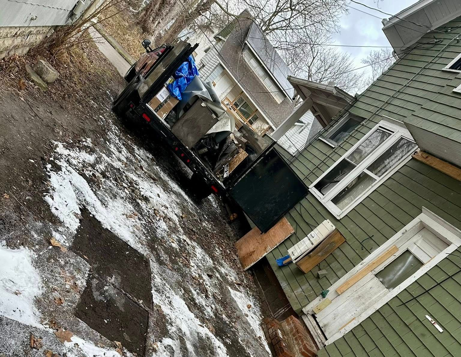 Tilted view of a snowy backyard with a green house, black trailer, and stacked items near the porch