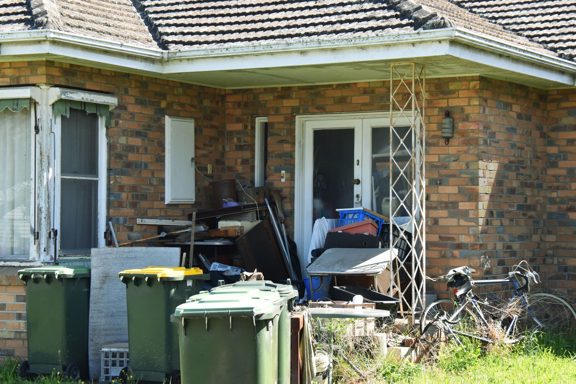 Front porch cluttered with bins, bikes, and household items outside a brick house.