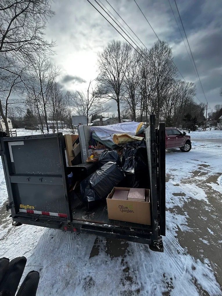 Overfilled dumpster trailer on snowy roadside with trash bags and boxes, car parked nearby