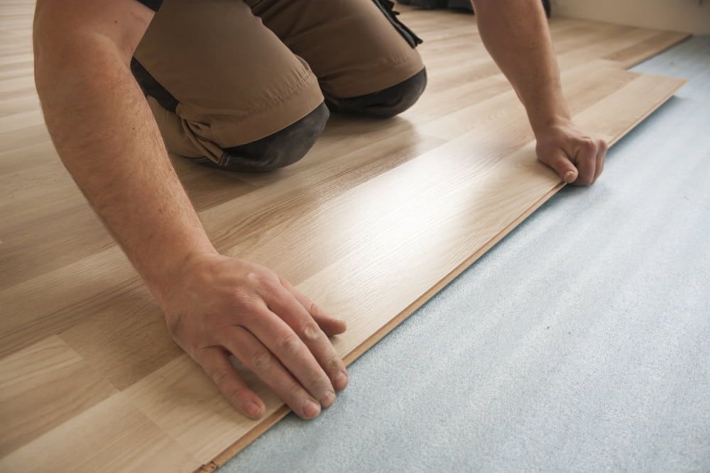 Person installing wood flooring; hands adjusting a plank over blue underlayment.