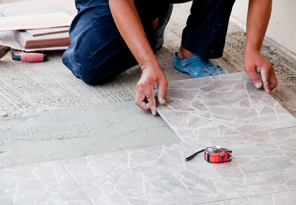 A Man Is Kneeling Down To Install A Tile Floor — Norm Evans Floors Pty Ltd in Beerwah, QLD