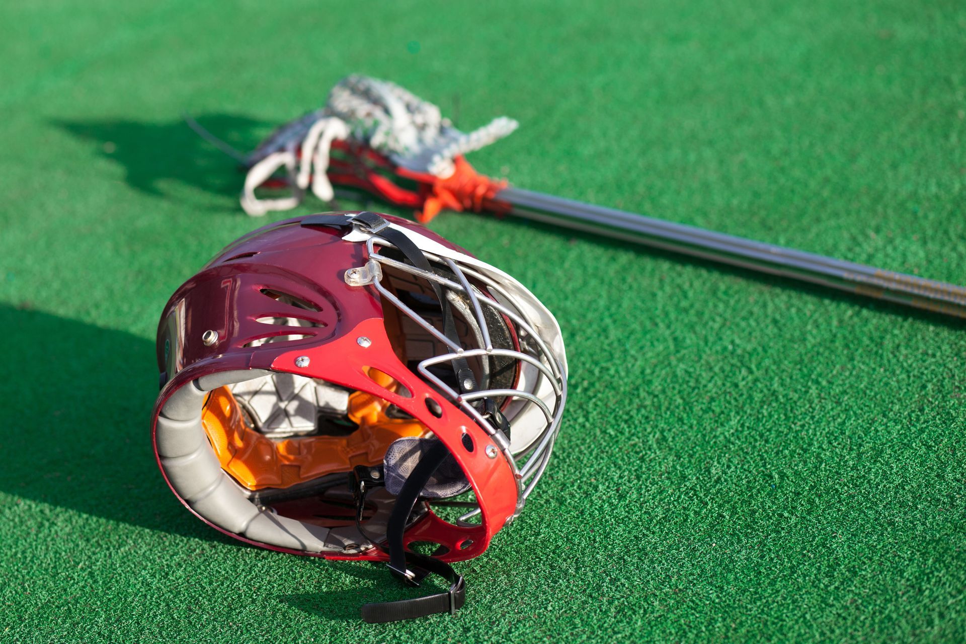 Lacrosse helmet with stick on green turf, maroon and black with orange chinstrap.