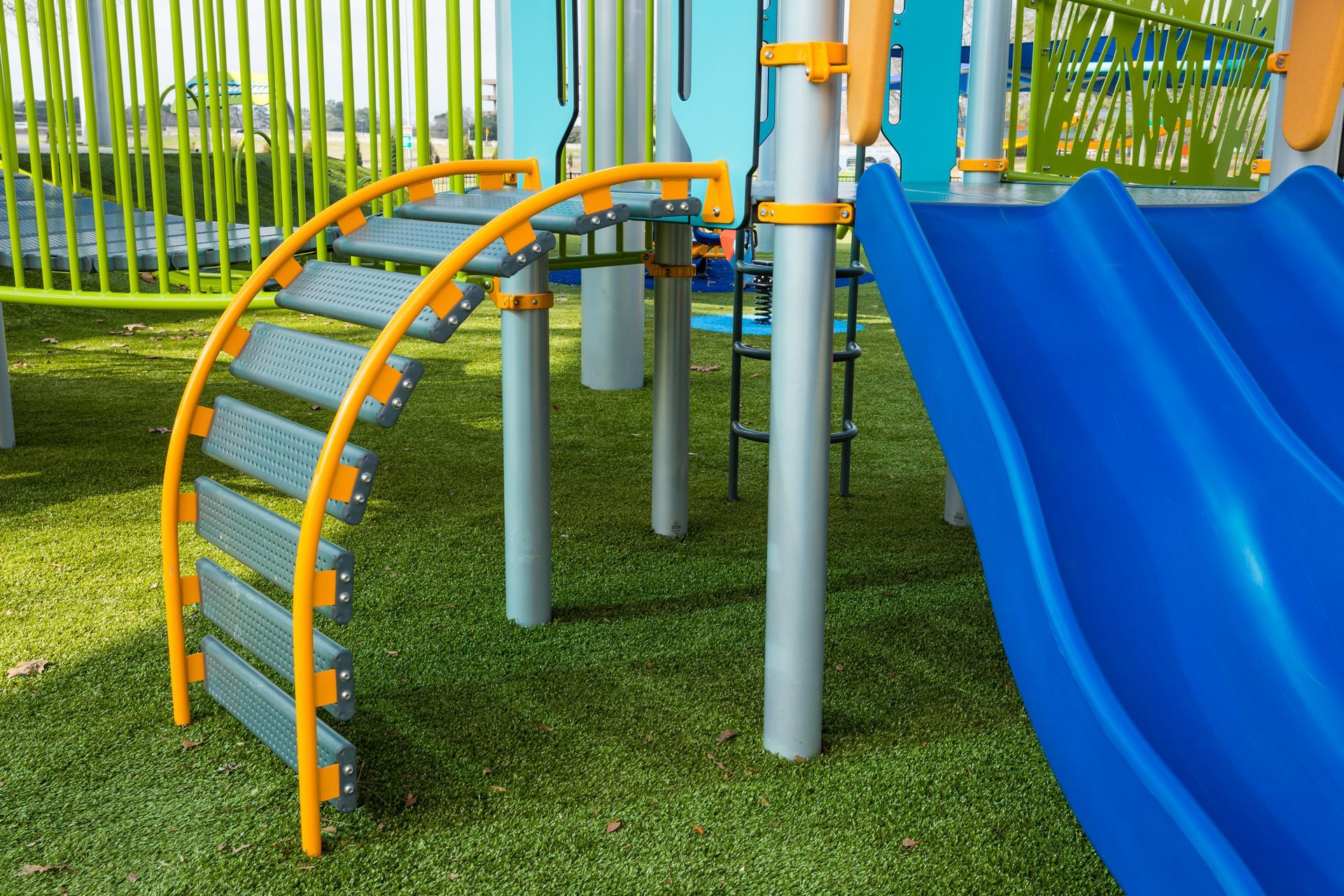 Colorful playground climbing arch and blue slide over grass with green fence in the background