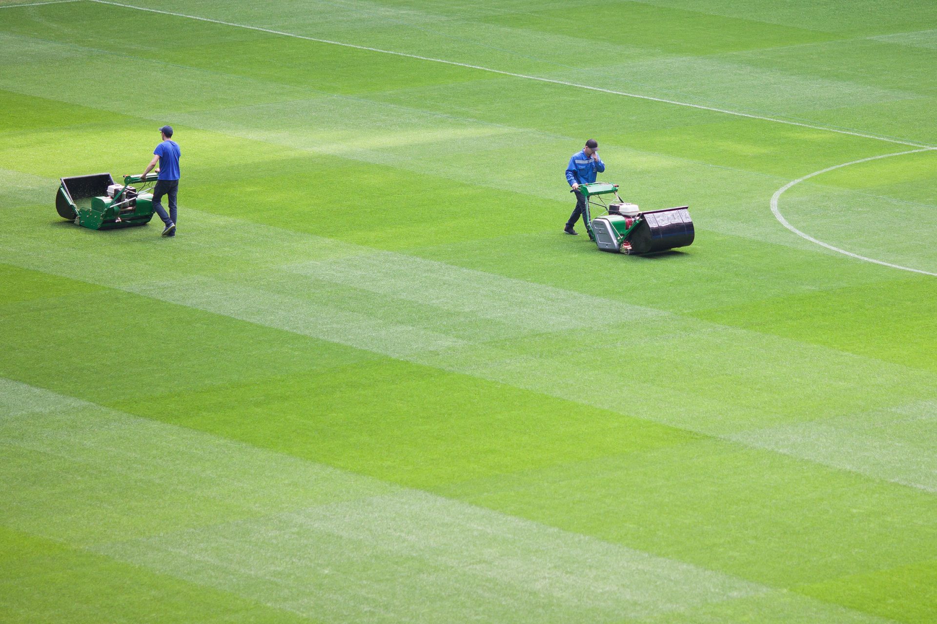 Two groundskeepers mowing a bright green sports field with ride-on mowers
